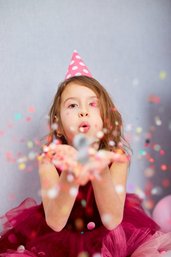 Beautiful little girl blows up multicolored confetti, having fun at home birthday party. Happy birthday child.
